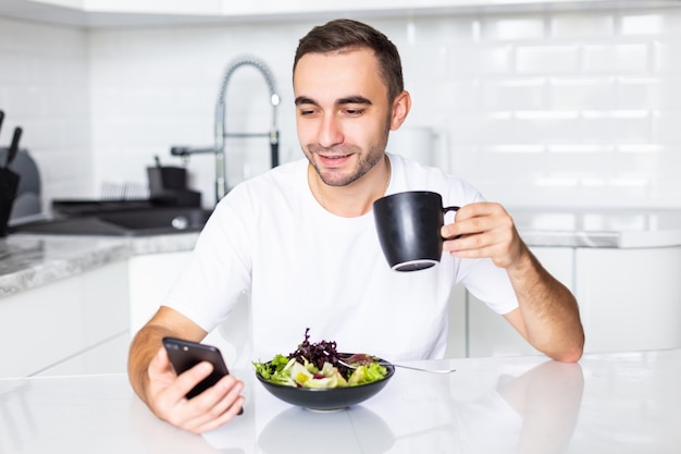 Hombre joven atractivo enviando mensajes de texto en su teléfono inteligente mientras desayuna en casa