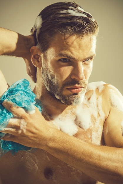 Hombre joven atractivo muscular que toma la ducha hombre desnudo frotándose el hombre de esponja de espuma con barba es