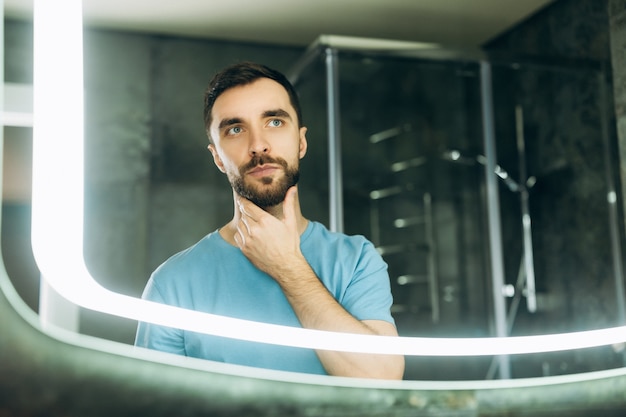 Hombre joven atractivo con ojos azules tocando su barba frente al espejo en la mañana