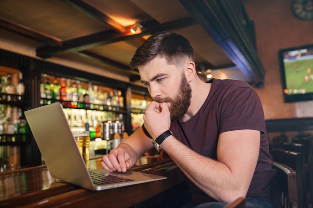 Hombre joven atractivo pensativo sentado y usando la computadora portátil en la barra