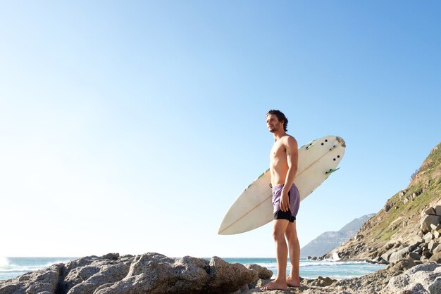 Hombre joven atractivo que se coloca en la playa que lleva la tabla hawaiana