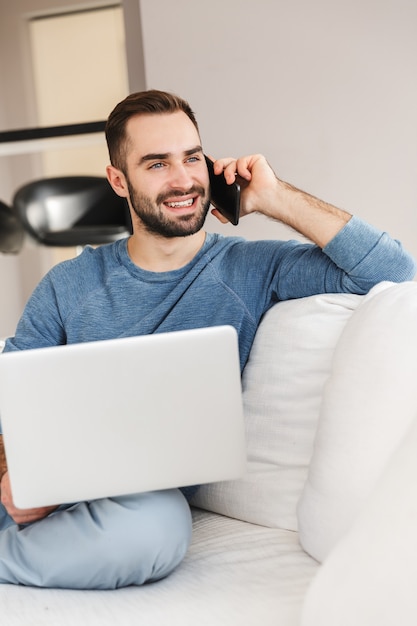 Hombre joven atractivo que se relaja en un sofá en casa, trabajando en la computadora portátil, hablando por teléfono móvil