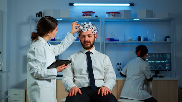 Hombre joven con auriculares de exploración de ondas cerebrales discutiendo con un investigador neurológico sobre los síntomas de la enfermedad. Científico médico escribiendo información de escaneo cerebral en tableta trabajando en laboratorio