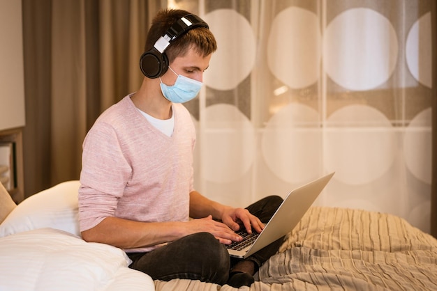 Foto hombre joven con auriculares y máscara protectora en casa con una computadora portátil