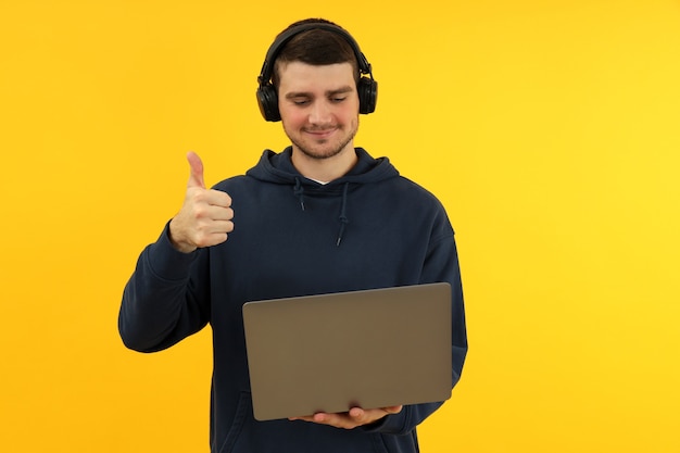 Hombre joven en auriculares sostiene la computadora portátil y muestra los pulgares para arriba sobre fondo amarillo.