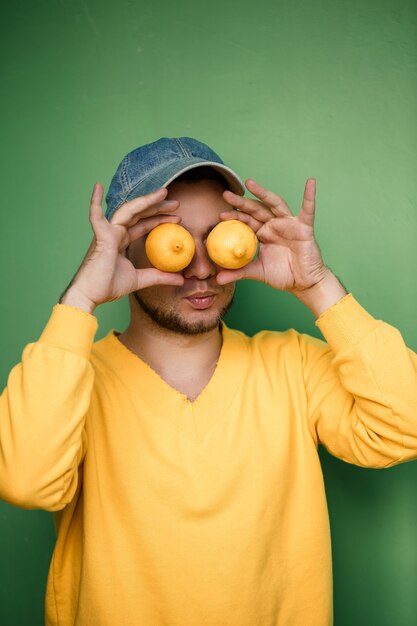 Hombre joven con barba en una gorra en la cabeza con limones en los ojos sobre un fondo verde. Retrato de un chico con un suéter amarillo