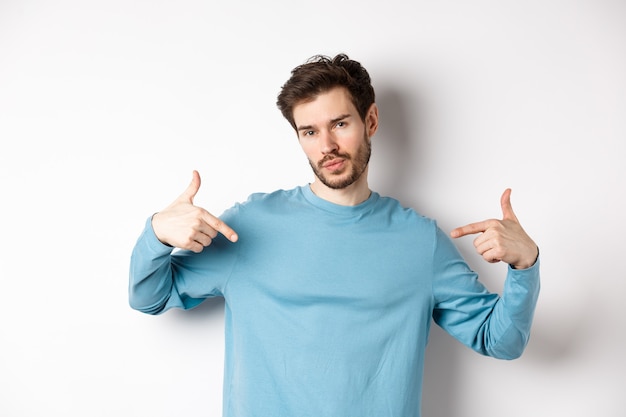 Hombre joven con barba que se siente confiado, apuntando a sí mismo y haciendo una cara engreída, de pie sobre un fondo blanco fresco y atrevido