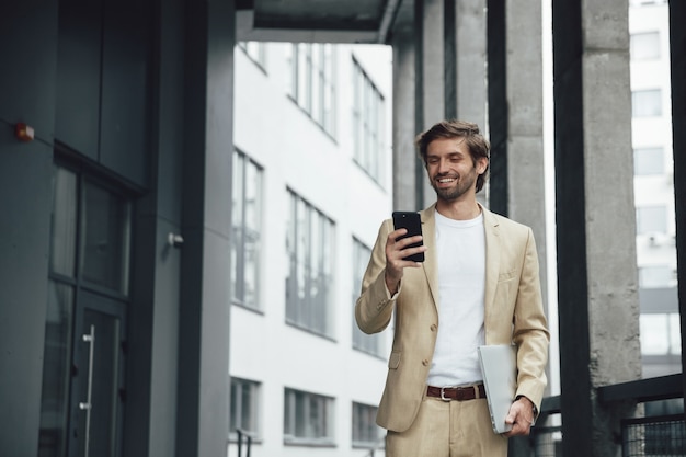 Hombre joven con barba en ropa formal sintiendo felicidad mientras mira la pantalla del teléfono inteligente. Empresario llevar portátil en la calle