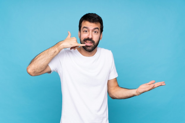 Hombre joven con barba sobre azul aislado haciendo gesto de teléfono y dudando