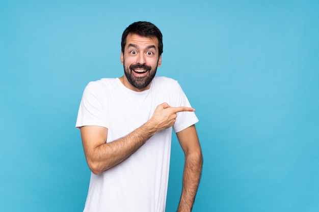 Hombre joven con barba sobre azul aislado sorprendido y apuntando con el dedo al lado