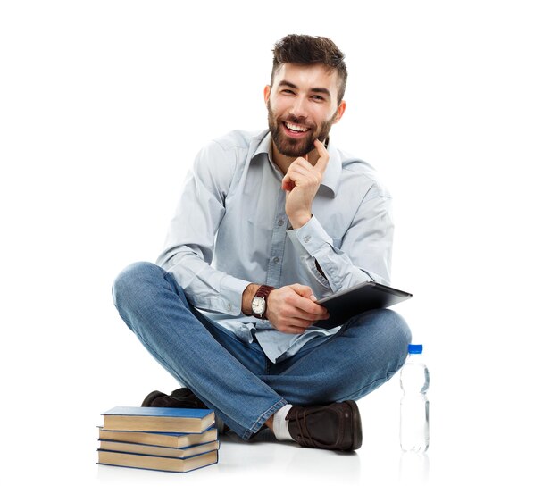 Hombre joven con barba sonriente sosteniendo una tableta con libros y una botella de agua sentado sobre un fondo blanco