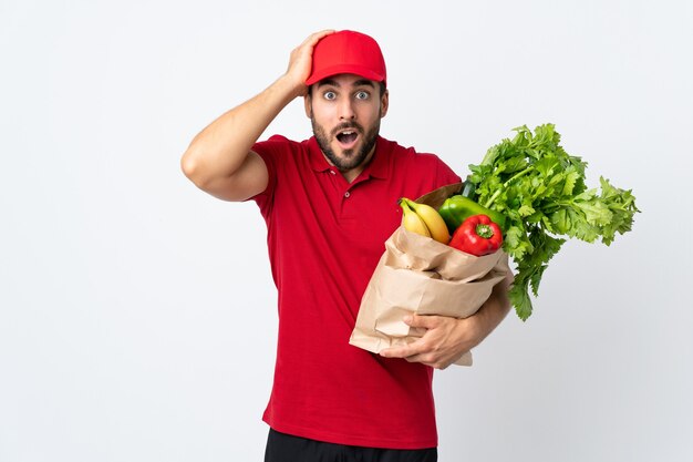 Hombre joven con barba sosteniendo una bolsa llena de verduras aislado en la pared blanca con expresión facial sorpresa