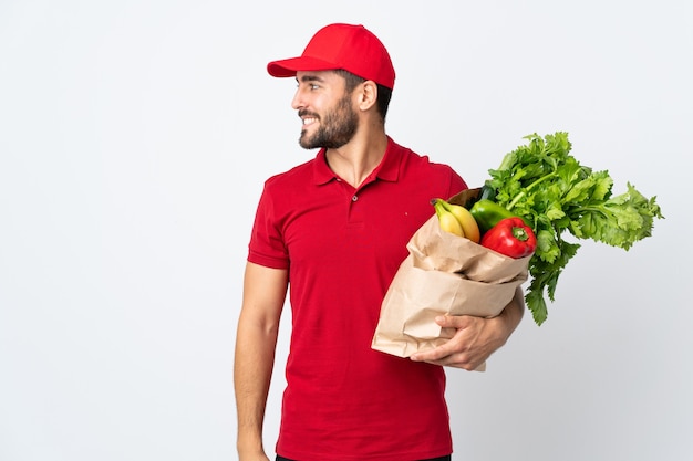 Hombre joven con barba sosteniendo una bolsa llena de verduras aislado en la pared blanca mirando hacia el lado