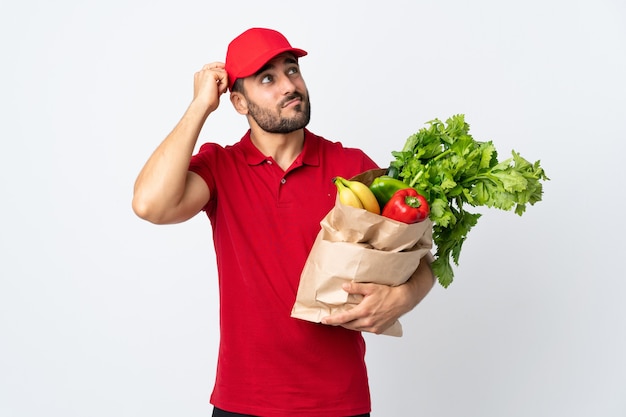 Hombre joven con barba sosteniendo una bolsa llena de verduras aislado sobre fondo blanco con dudas y con expresión de la cara confusa