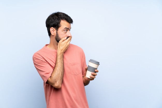 Hombre joven con barba sosteniendo un café para llevar sobre azul aislado cubriendo la boca y mirando hacia el lado