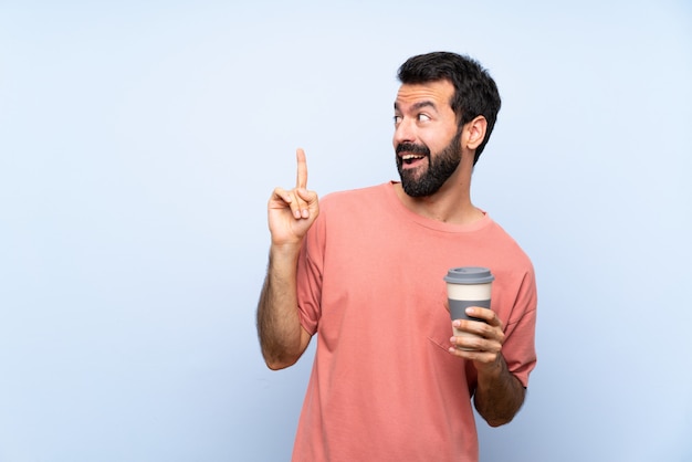 Hombre joven con barba sosteniendo un café para llevar sobre una pared azul aislada con la intención de darse cuenta de la solución mientras levanta un dedo