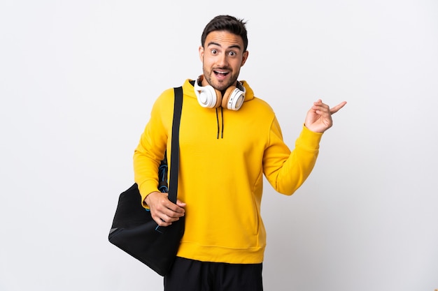 Hombre joven con bolsa de deporte aislado en blanco sorprendido y apuntando con el dedo hacia el lado