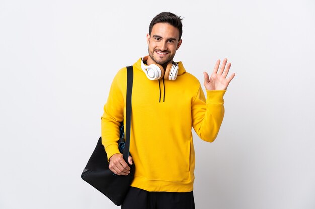 Hombre joven con bolsa de deporte aislado en la pared blanca saludando con la mano con expresión feliz