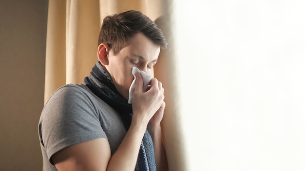Hombre joven con bufanda caliente manteniendo los ojos cerrados y sonándose la nariz con un pañuelo de papel mientras se siente mal y de pie cerca de la ventana en casa, copie el espacio