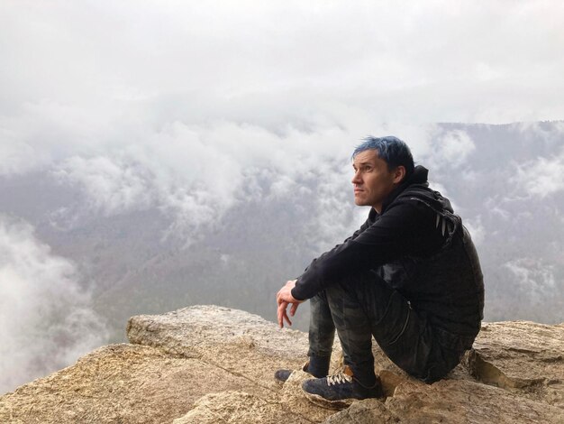 Hombre joven con cabello azul sentado en el borde del acantilado en un clima nublado y brumoso Turista masculino disfruta de una vista espectacular de la naturaleza sentado en una roca alta