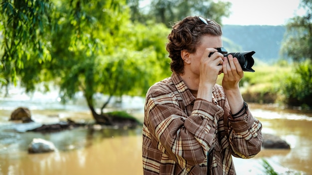 Un hombre joven con cabello rizado tomando fotografías con la cámara en la naturaleza, el río y la vegetación cercana