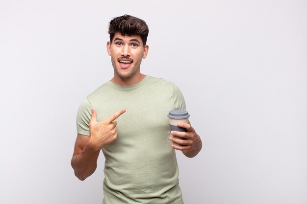 Hombre joven con un café mirando emocionado y sorprendido apuntando hacia un lado y hacia arriba para copiar el espacio