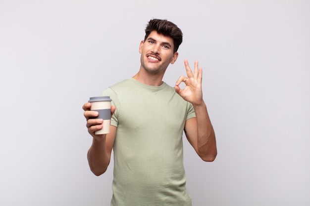 Hombre joven con un café sintiéndose feliz, relajado y satisfecho, mostrando aprobación con gesto bien, sonriendo
