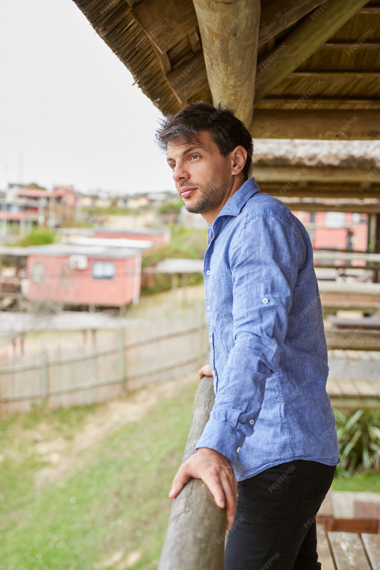 Hombre joven con una camisa azul claro en la barandilla de madera de la terraza de su cabaña mira horizonte. | Foto Premium