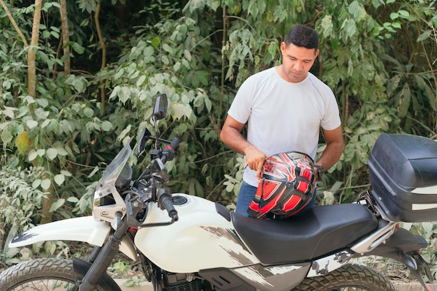 Hombre joven con camisa blanca y jeans poniéndose el casco cerca de la motocicleta al aire libre