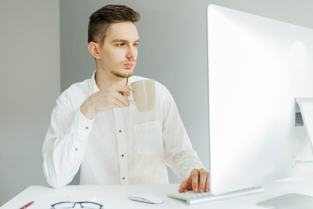 Hombre joven en camisa blanca con taza de trabajo en el escritorio con una computadora en la oficina. Trabajador sobre fondo gris