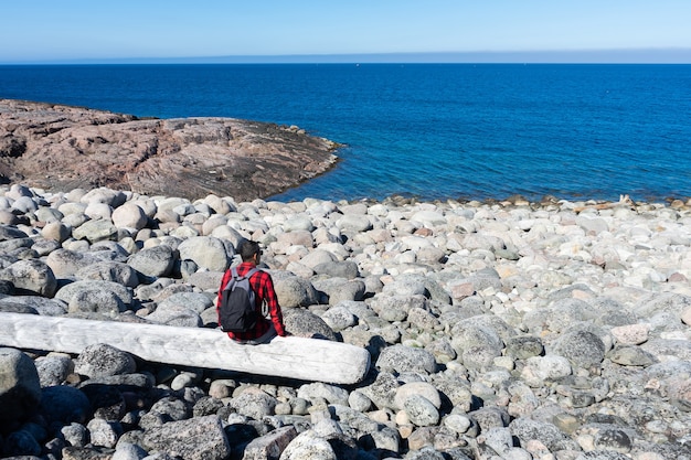 Un hombre joven con una camisa a cuadros roja se sienta en un tronco solo en la orilla del mar