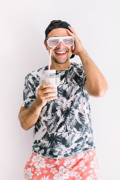 Hombre joven con una camisa floral, gorra en verano, bebiendo un cóctel con una pajita y haciendo gestos alegres, a la luz del día en una pared blanca