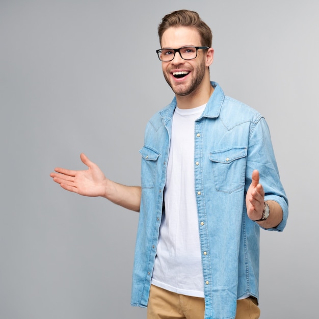 Hombre joven con camisa de jeans dándole la bienvenida con una sonrisa en su rostro y sus brazos abiertos de pie sobre la pared gris