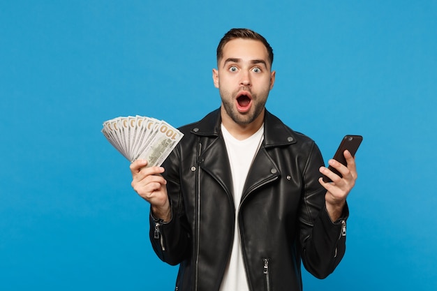 Hombre joven en camiseta blanca de chaqueta de cuero negro con ventilador de dinero en efectivo en billetes de dólar, teléfono celular aislado en retrato de estudio de fondo de pared azul. Concepto de estilo de vida de personas. Simulacros de espacio de copia.