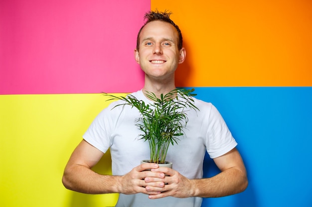 Hombre joven en una camiseta blanca, fondo colorido, palma decorativa, foto de emociones, planta casera, amor por la naturaleza
