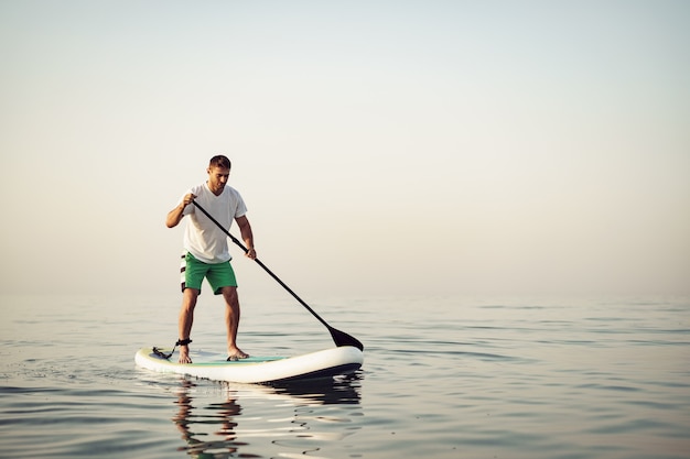 Hombre joven en camiseta y pantalones cortos flotando a bordo de sup