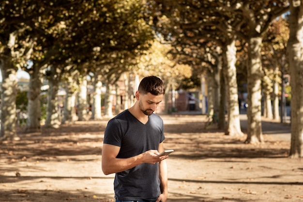 Hombre joven en camiseta de pie en la calle con teléfono móvil y escribiendo mensajes de texto.