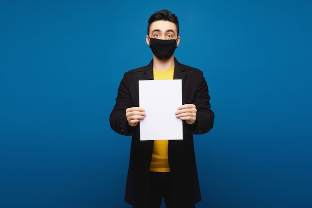 Hombre joven en una capa negra y una máscara protectora negra que presenta con una hoja de papel vacía en el fondo azul, concepto de la promoción. Concepto de salud