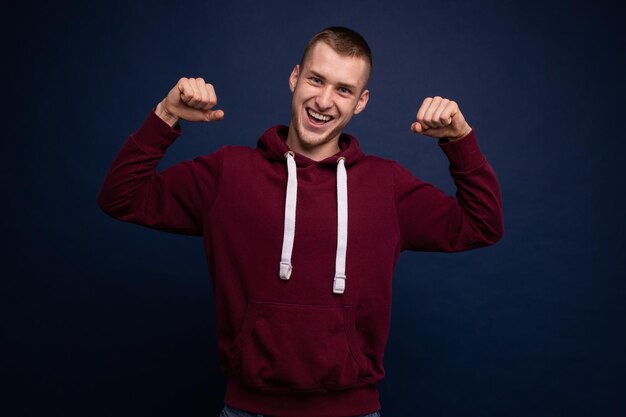 Hombre joven con capucha roja y jeans posando sobre fondo azul sonriendo y mostrando buena suerte