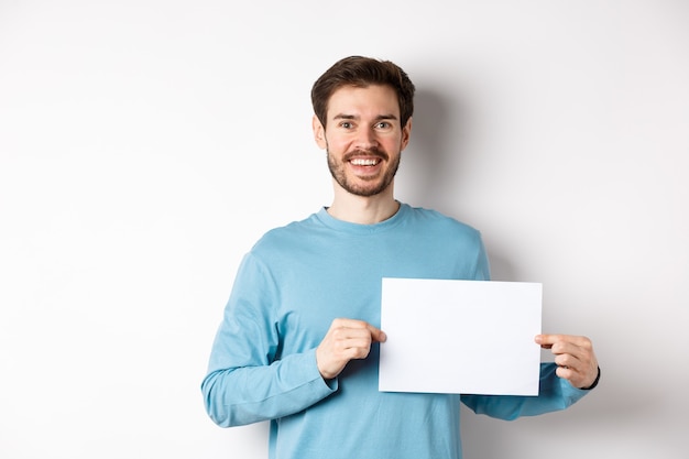 Hombre joven con cara feliz mostrando un trozo de papel en blanco para su logotipo o cartel, sonriendo a la cámara, de pie sobre fondo blanco.