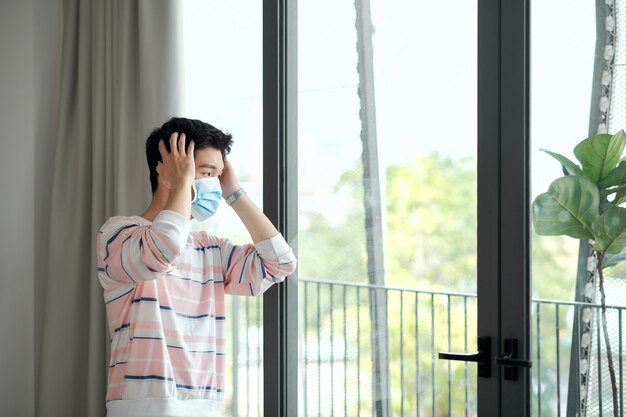 Hombre joven en casa en cuarentena con máscara médica junto a la ventana de fondo blanco. Coronavirus, enfermedad, infección, cuarentena, vendaje quirúrgico, máscara médica, depresión