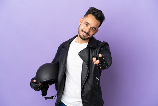 Hombre joven con un casco de motocicleta aislado sobre fondo púrpura apuntando hacia el frente con expresión feliz