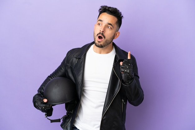 Hombre joven con un casco de motocicleta aislado sobre fondo púrpura pensando en una idea apuntando con el dedo hacia arriba