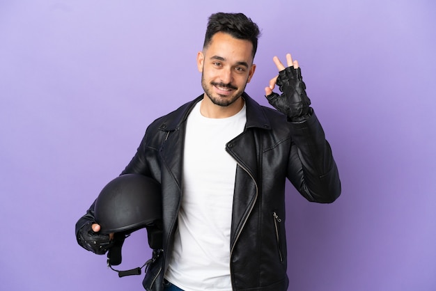 Hombre joven con un casco de motocicleta aislado sobre fondo púrpura que muestra el signo de ok con los dedos