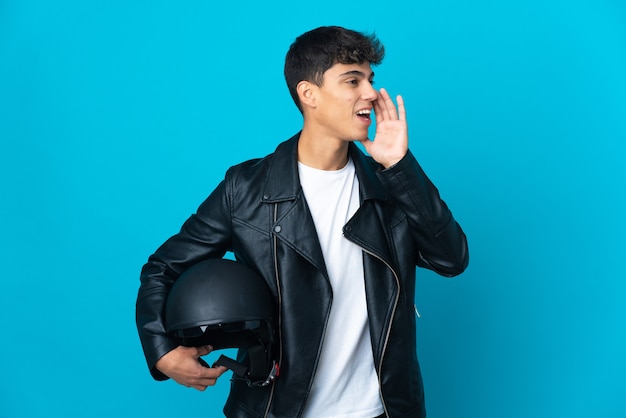 Hombre joven con un casco de motocicleta sobre pared azul aislada gritando con la boca abierta hacia un lado