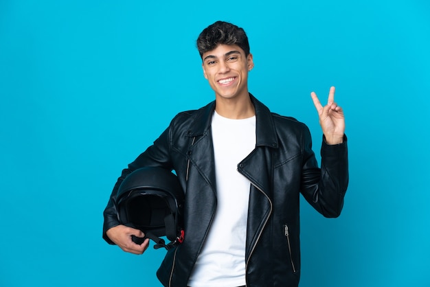 Hombre joven con un casco de motocicleta sobre pared azul aislada sonriendo y mostrando el signo de la victoria