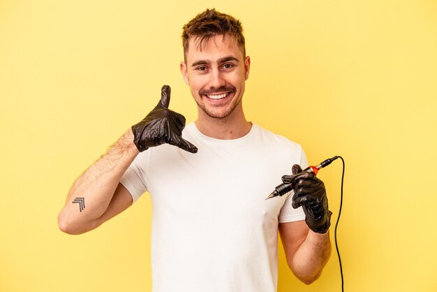 Hombre joven caucásico tatuador aislado sobre fondo amarillo que muestra un gesto de llamada de teléfono móvil con los dedos.