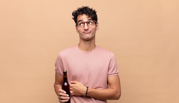 Hombre joven con una cerveza encogiéndose de hombros, sintiéndose confundido e inseguro, dudando con los brazos cruzados y la mirada perpleja