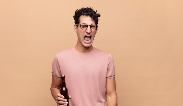 Hombre joven con una cerveza gritando agresivamente, luciendo muy enojado, frustrado, indignado o molesto, gritando no