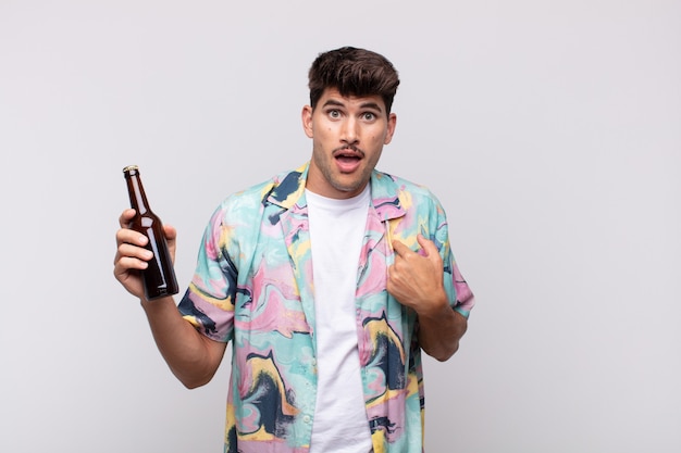 Hombre joven con una cerveza que se siente feliz, sorprendido y orgulloso, apuntando a sí mismo con una mirada emocionada y asombrada